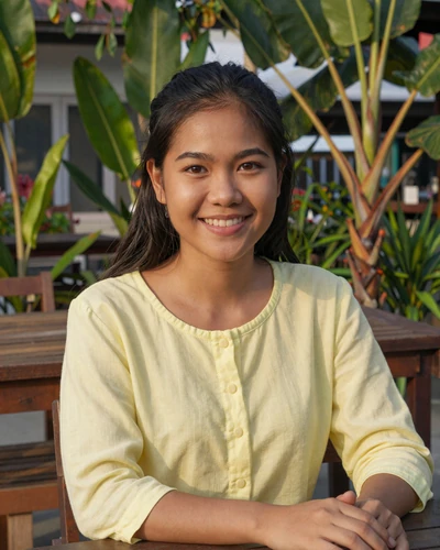 Nong, 24, Thai woman in yellow blouse smiling at outdoor cafe in Chiang Rai